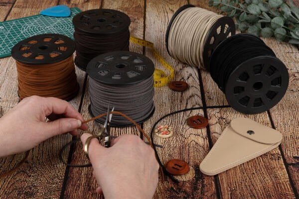 A person cutting brown braided leather cord with scissors. Spools of braided leather in various colors, a measuring tape, a ruler, buttons, a small leather pouch, and green plant leaves are arranged on a wooden surface.