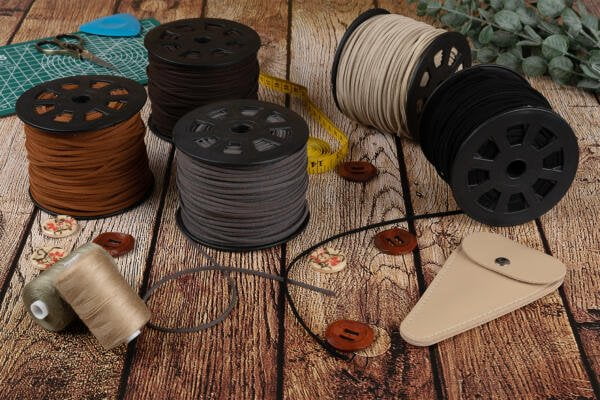 A rustic tabletop is scattered with various sewing supplies including several spools of colored threads, a measuring tape, buttons, scissors, a green cutting mat, green foliage, and a beige leather pouch. The surface is made of weathered wooden planks.