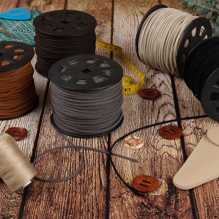 A rustic wooden table is filled with sewing supplies: several spools of brown, gray, and beige thread, a yellow measuring tape, brown buttons, a beige leather needle case, and decorative buttons with floral designs. A plant is partially visible in the corner.