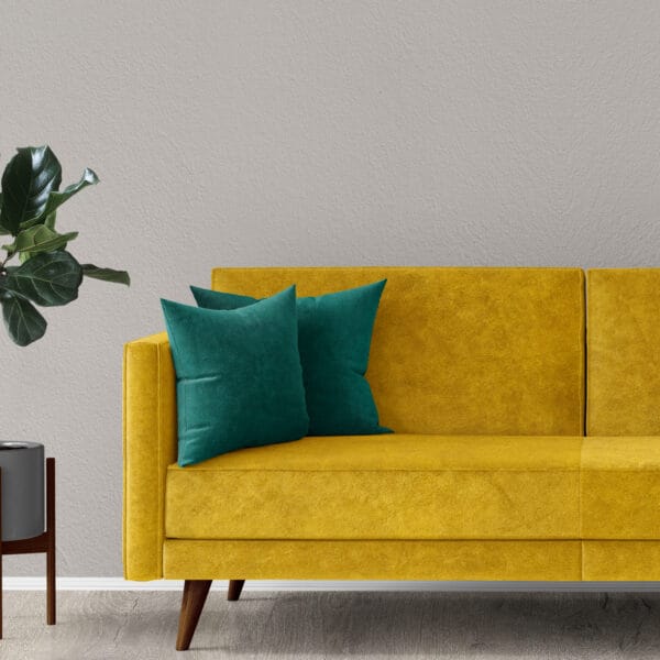A modern sofa upholstered in Velvet Fabric | Plush Super Velvet Fabric | 60 Inch Wide with wooden legs and teal pillows sits by a light gray wall, while a green potted plant on a wooden stand is placed to its left on a light floor.