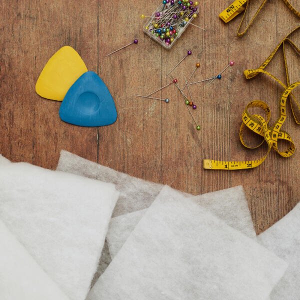 A wooden surface displays sewing tools, including yellow and blue tailor's chalk, a container of colorful pins, scattered pins, a yellow measuring tape, and several pieces of white fabric.