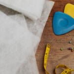 A close-up of sewing supplies on a wooden surface includes white fabric, colored tailor's chalk in blue and yellow, a yellow measuring tape, and assorted pins with colorful heads.