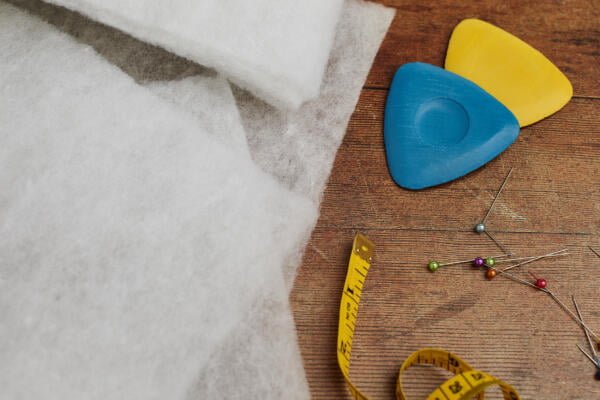 A close-up of sewing supplies on a wooden surface includes white fabric, colored tailor's chalk in blue and yellow, a yellow measuring tape, and assorted pins with colorful heads.