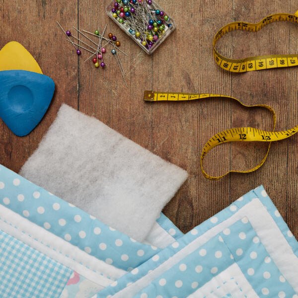 A quilting workspace on a wooden table featuring a measuring tape, colored pins in a clear box, blue tailor's chalk, a sponge pad, a pin cushion filled with white filling, and a blue and white polka dot quilt in progress.