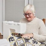 An older woman with short white hair and glasses is smiling while sewing a quilt on a Pfaff sewing machine. She is seated at a table, wearing a beige sweater, with spools of thread and pins nearby. The quilt has a black, white, and yellow pattern.