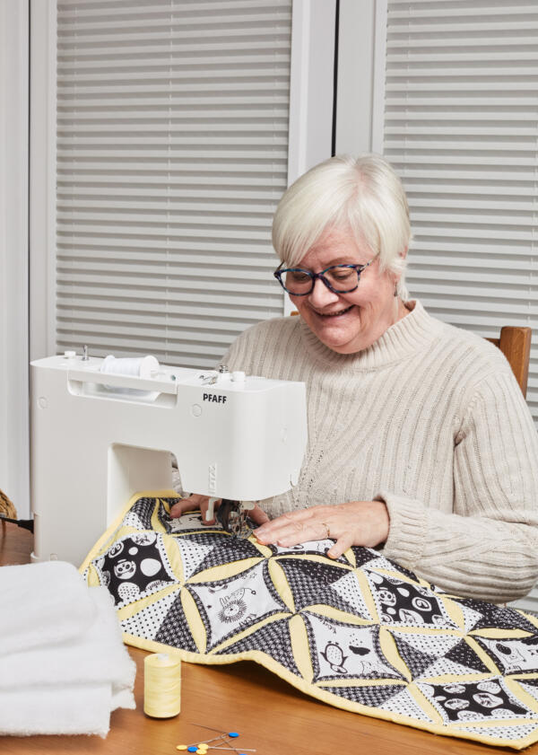 An older woman with short white hair and glasses is smiling while sewing a quilt on a Pfaff sewing machine. She is seated at a table, wearing a beige sweater, with spools of thread and pins nearby. The quilt has a black, white, and yellow pattern.