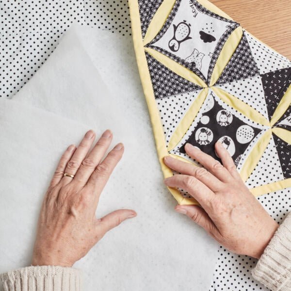 Close-up of an older adult's hands working on a quilt with a black and white pinwheel pattern and a yellow border. The quilt has cat and bird designs. The person is assembling the quilt with batting and fabric on a polka dot surface.
