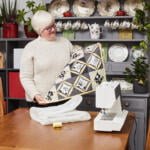 An elderly woman with short white hair and glasses is standing by a table with a sewing machine. She is holding a quilted blanket with a black, white, and yellow pattern. Shelves with plates, teacups, and plants are in the background. The mood is cozy and creative.