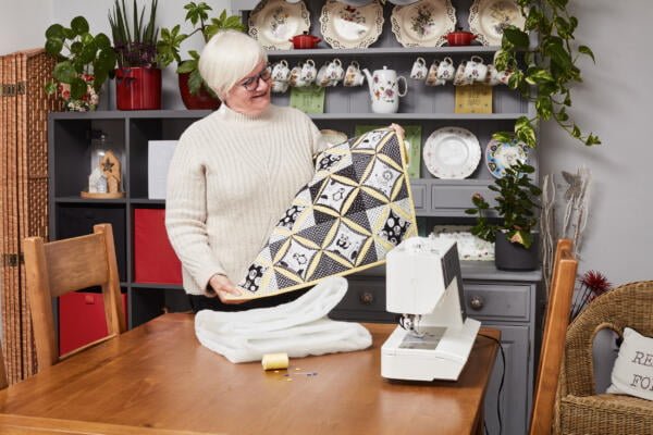 An elderly woman with short white hair and glasses is standing by a table with a sewing machine. She is holding a quilted blanket with a black, white, and yellow pattern. Shelves with plates, teacups, and plants are in the background. The mood is cozy and creative.