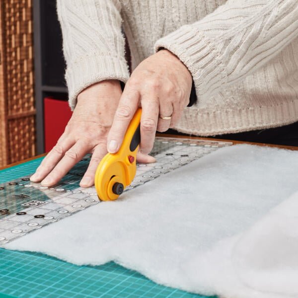 A person wearing a white sweater is using a yellow rotary cutter and a metal ruler on a green cutting mat to cut a piece of white fabric or batting. The rotary cutter is held in the right hand, guiding along the ruler, while the left hand holds the ruler steady.