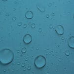 A close-up of the "Waterproof Fabric | Waterproof Canvas | 60 Inches Wide" in blue reveals a textured surface with scattered water droplets, giving a fresh look. The droplets reflect light, creating a shimmering effect.