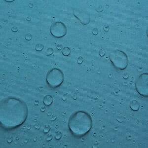 A close-up of the "Waterproof Fabric | Waterproof Canvas | 60 Inches Wide" in blue reveals a textured surface with scattered water droplets, giving a fresh look. The droplets reflect light, creating a shimmering effect.