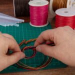 Two hands are weaving colorful cords on a green cutting mat, surrounded by various spools of thread and some glue sticks on a wooden table. The cords include red, yellow, and black colors, and the person appears to be making a bracelet or similar craft item.