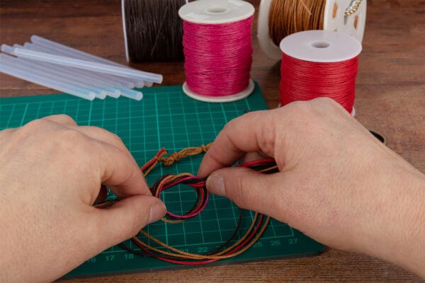 Two hands are weaving colorful cords on a green cutting mat, surrounded by various spools of thread and some glue sticks on a wooden table. The cords include red, yellow, and black colors, and the person appears to be making a bracelet or similar craft item.
