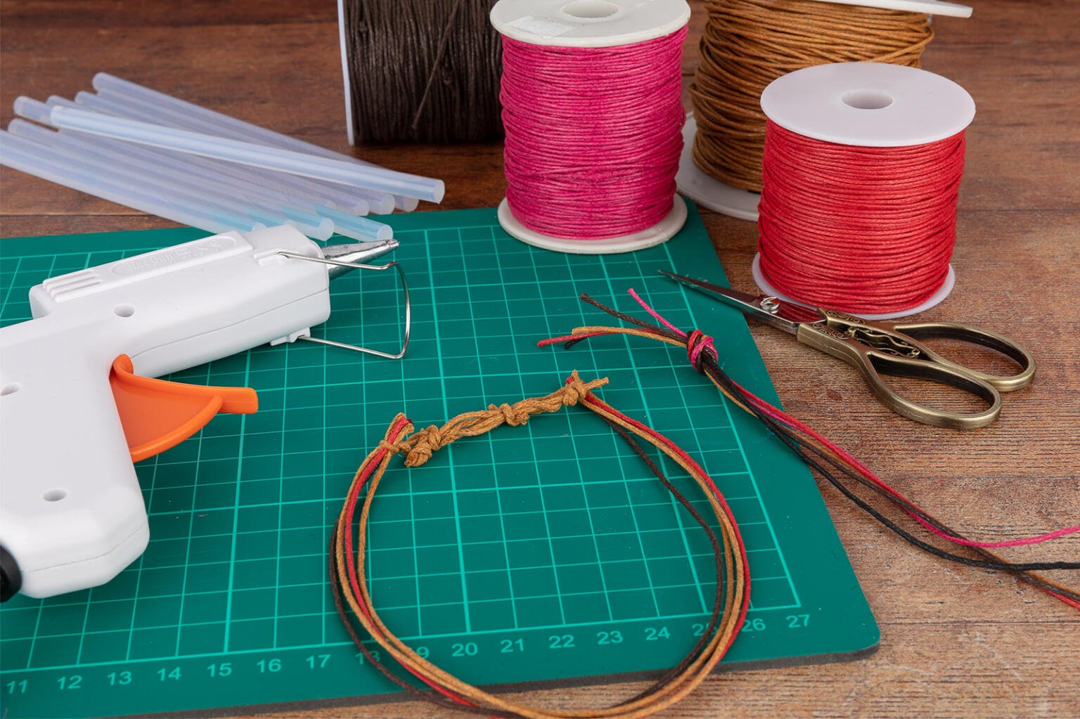 A green cutting mat with crafting supplies, including a white glue gun, glue sticks, spools of pink, red, and brown cord, scissors, and a partially crafted bracelet made of multicolored cords, is placed on a wooden surface.