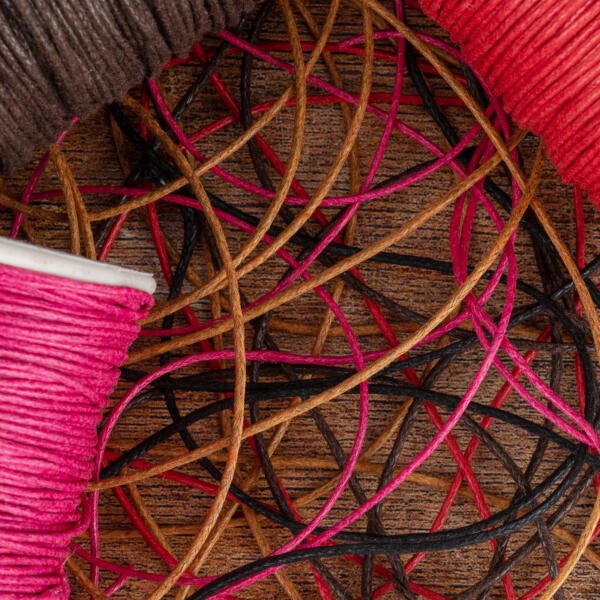 Colorful spools of yarn, including pink, brown, black, and orange, are intertwined on a wooden surface, creating a tangled pattern of threads. The various hues and textures add a vibrant and chaotic visual interest to the scene.