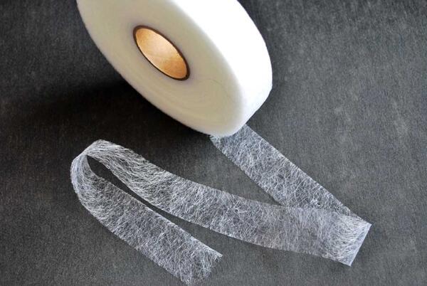 A roll of lightweight white interfacing tape is unwound on a dark surface. The translucent material appears textured and is used in sewing and crafting for reinforcing fabric.