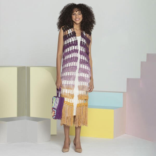 A woman with curly hair is wearing a long, sleeveless, multicolored crochet vest with fringe, paired with a white dress and beige heels. She is carrying a colorful woven bag. The background features pastel-colored geometric shapes.