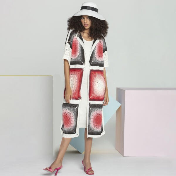 A woman with curly hair, wearing a white wide-brimmed hat, multi-colored long cardigan with red and black patterns, white dress underneath, and pink heels, stands confidently against a minimalist background with pastel geometric shapes.