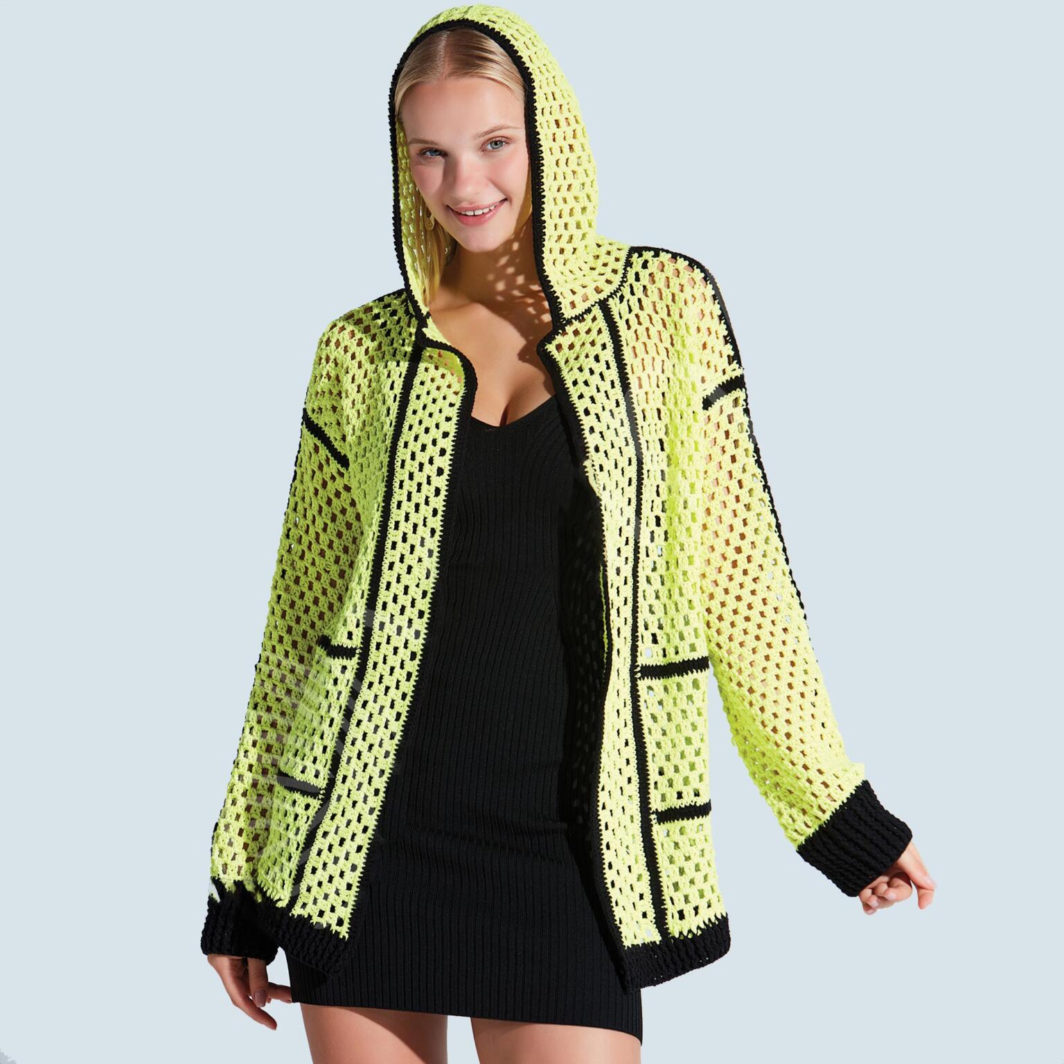 A woman wearing a bright yellow, see-through mesh hoodie with black trim over a black dress stands smiling against a light blue background.