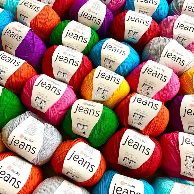 A neatly stacked collection of YarnArt Jeans yarn balls from PAC Yarn showcases a vibrant assortment, featuring shades of red, blue, green, orange, and purple for a visually appealing and organized display.