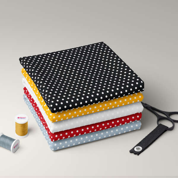 A neatly stacked pile of folded Cotton Poplin | Polka Dot | Rose and Hubble fabrics in black, yellow, white, red, and light blue colors. Nearby are a spool of yellow thread, a spool of light blue thread, a pair of scissors, and a measuring tape. The background is a smooth, light-colored surface.