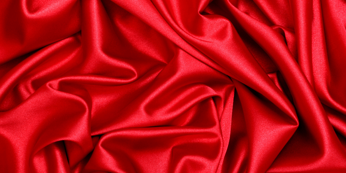A close-up view of luxurious red silk fabric arranged in soft, flowing folds and creases. The material appears smooth and shiny, reflecting light subtly across its surface, highlighting its rich color and texture.