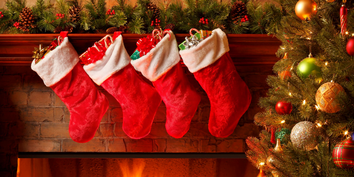 Four red Christmas stockings hang from a decorated mantel above a fireplace. Each stocking is filled with wrapped gifts. The mantel is adorned with green garlands and red ornaments. A lit Christmas tree with colorful ornaments stands to the right.