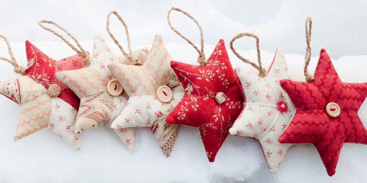 A row of eight star-shaped fabric ornaments in various red and white patterns hang by jute strings. These charming stars, each with a button at the center, are displayed against a snowy background – a perfect Christmas idea for festive decor.