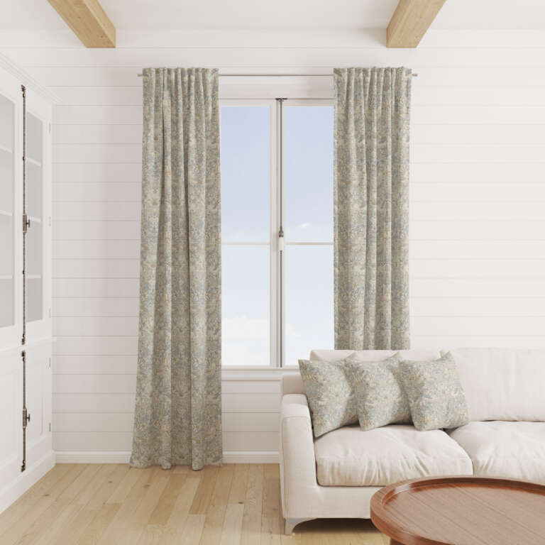 A cozy living room with a white shiplap wall, a large window with light blue patterned curtains, a white couch adorned with matching patterned throw pillows, wooden floor, and a round wooden coffee table in the foreground.