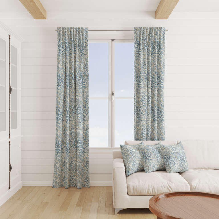 A cozy, bright living room with a white couch adorned with patterned blue and white pillows. A large window with patterned blue and white curtains lets in ample natural light. The room features wooden beams on the ceiling and a wooden floor, creating a warm ambiance.