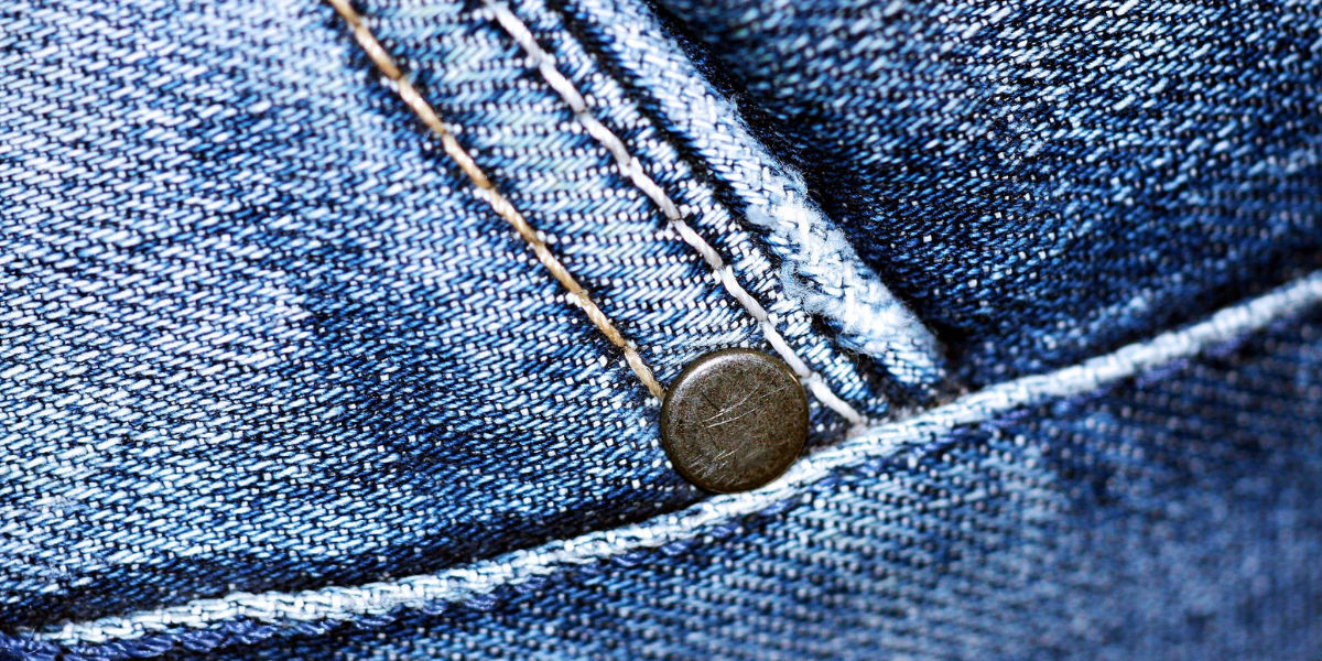 Close-up of blue denim fabric showing detailed stitching and a metal rivet. The image highlights the texture and pattern of the fabric, with visible threads and seams.