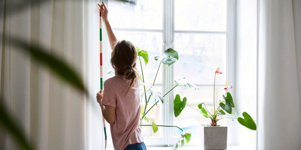 A person in a pink shirt hangs a colorful striped decoration on a white curtain next to a window. The windowsill features green potted plants, and sunlight streams through the window, creating a bright and airy atmosphere in the room.