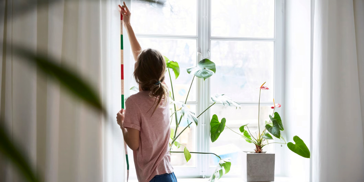 A person in a pink shirt hangs a colorful striped decoration on a white curtain next to a window. The windowsill features green potted plants, and sunlight streams through the window, creating a bright and airy atmosphere in the room.