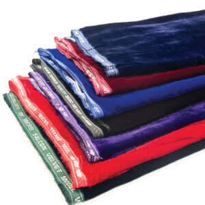 A neatly stacked assortment of velvet fabric in various colors, including blue, red, green, purple, and black, each with branded selvedge edges. The fabrics have a soft and shiny texture, showcasing the rich and lustrous quality of the velvet material.