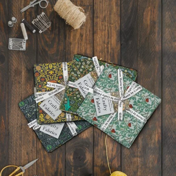 A wooden table holds six fabric bundles from the "William Morris Christmas Fat Quarter Bundle" collection, featuring intricate floral patterns in various colors, all neatly tied with white ribbons labeled "Crafty Fabrics." Surrounding them are sewing tools, twine, a yellow measuring tape, and a filled pincushion.