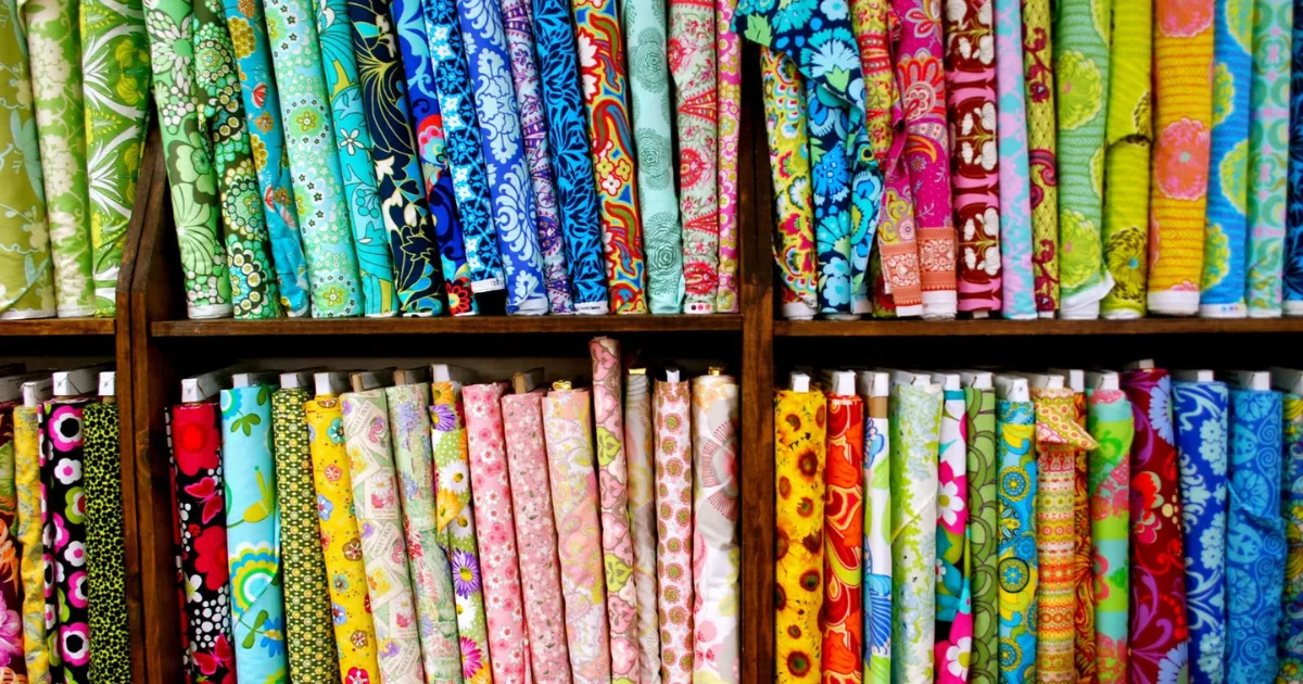 A colorful assortment of fabric rolls are neatly arranged on shelves. The fabrics feature various patterns, including floral designs, geometric shapes, and abstract prints, in a wide range of vibrant colors such as blue, pink, green, and yellow.