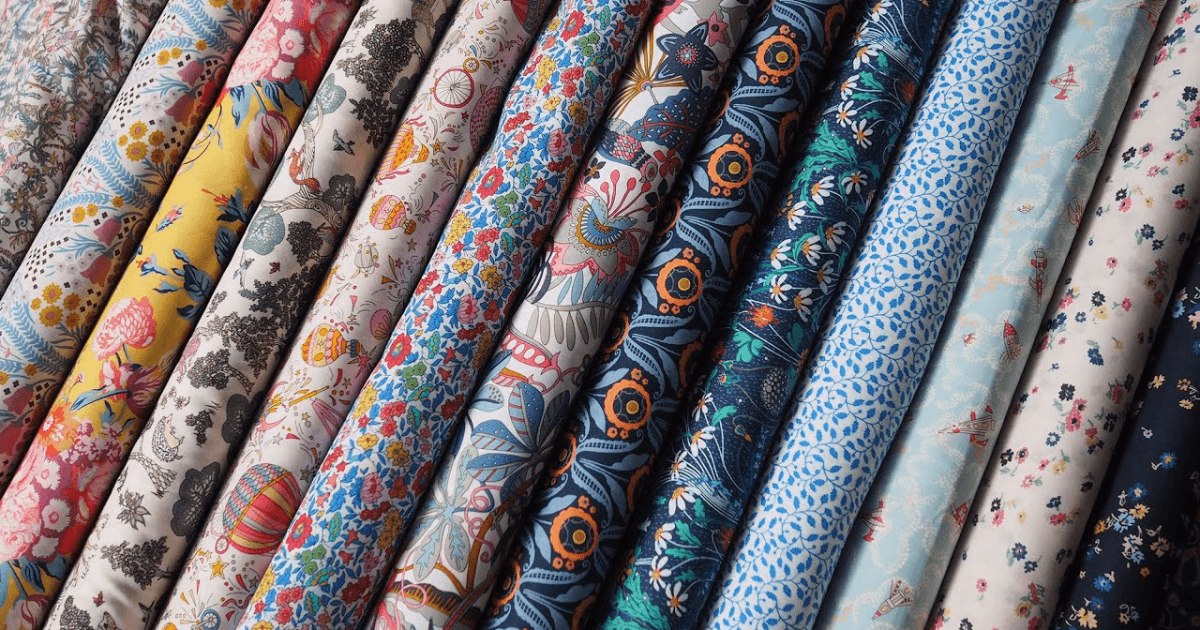 A variety of colorful fabric rolls displayed vertically, each featuring distinct floral and geometric patterns. The vibrant patterns include florals, abstract shapes, and intricate designs, creating a diverse and visually appealing assortment.