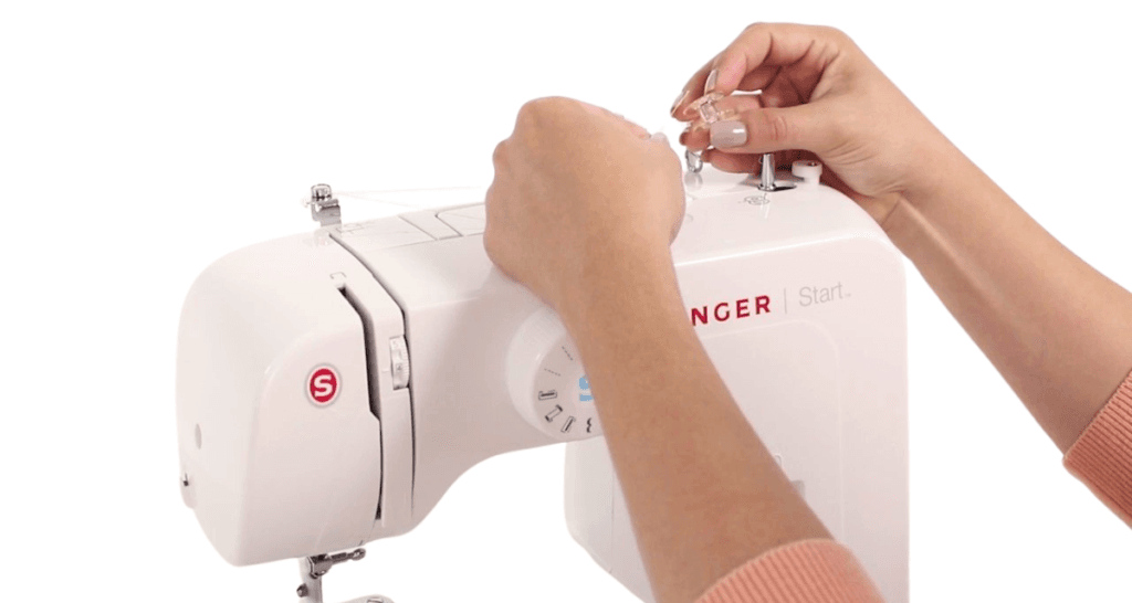 Close-up of hands threading a Singer sewing machine, with one hand holding the thread and the other skillfully guiding it through the sewing machine's components. The white machine stands out against a plain background.