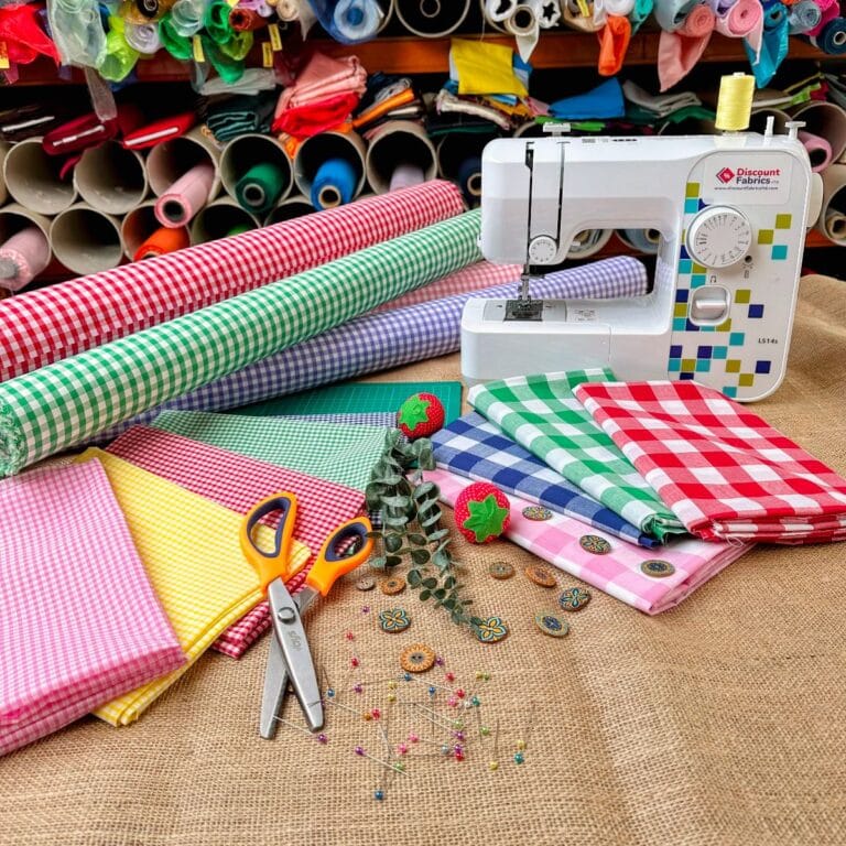 A sewing machine on a fabric-covered table surrounded by colorful rolls and pieces of gingham fabric. Two pairs of scissors, sewing accessories, and decorative buttons are nearby, with spools of thread on shelves in the background.