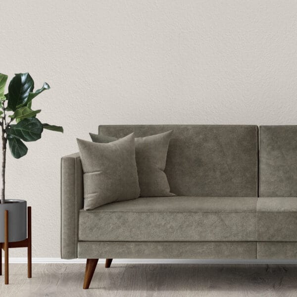 A modern living room features a minimalist green velvet sofa with two matching cushions. A potted green plant in a gray pot stands beside it, set against an off-white textured wall and light wood flooring.