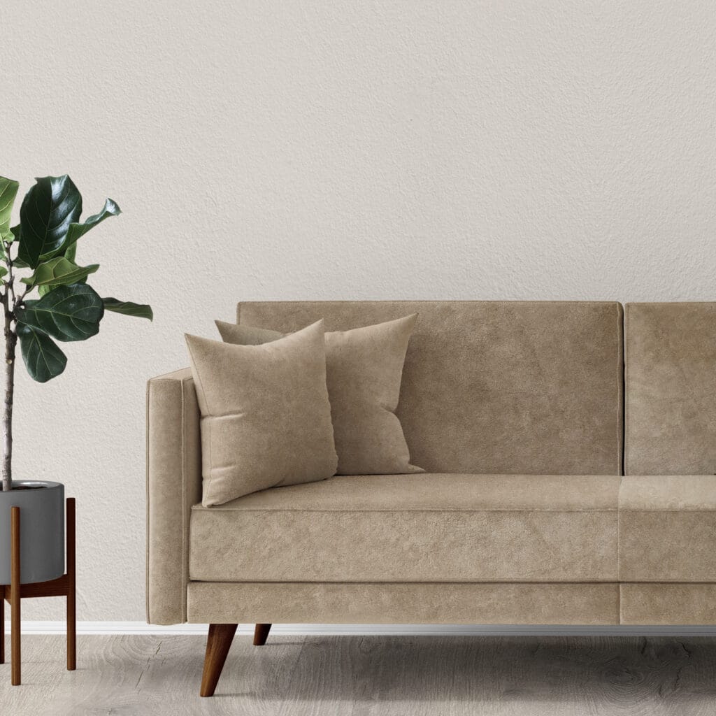 A beige suede sofa with two matching cushions is placed against a light-colored textured wall. To the left, there's a potted plant with broad green leaves in a gray planter on a wooden stand. The floor is light wood.