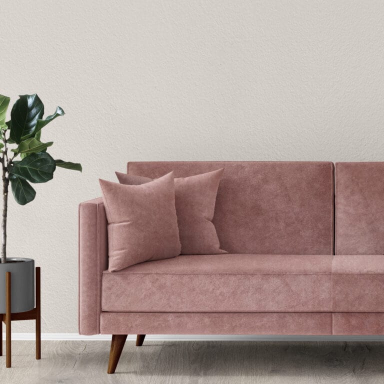 A modern, minimalist living room features a pink sofa with two matching cushions, a potted green plant in a sleek gray planter, and a light gray textured wall in the background. The floor is light wood, complementing the neutral tones.