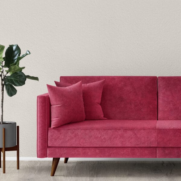 A modern living room features a plush pink sofa with wooden legs and matching throw pillows against a light gray wall. A potted plant with broad green leaves stands to the side on a wooden floor.