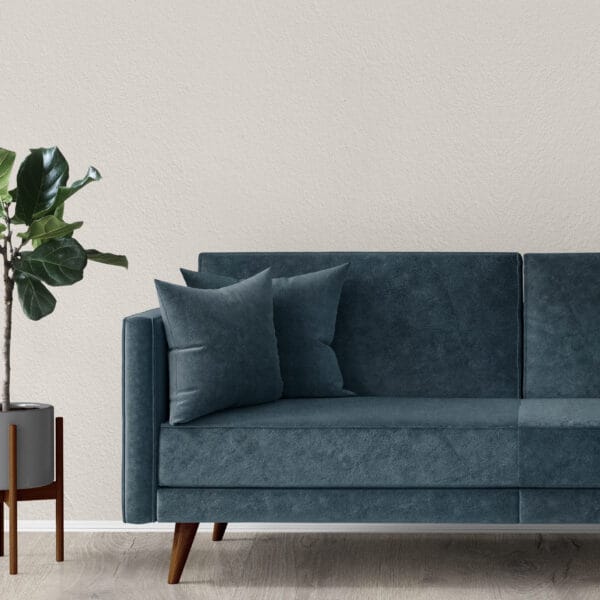 A modern living room features a dark teal velvet sofa with wooden legs, adorned with matching throw pillows. Next to the sofa is a tall potted plant in a sleek planter. The background is a textured white wall and light wooden floor.
