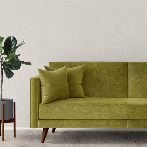 A modern living room with a green velvet sofa adorned with two matching cushions. The sofa stands on light wooden flooring, next to a tall potted plant with a gray pot, set against a plain, off-white wall.