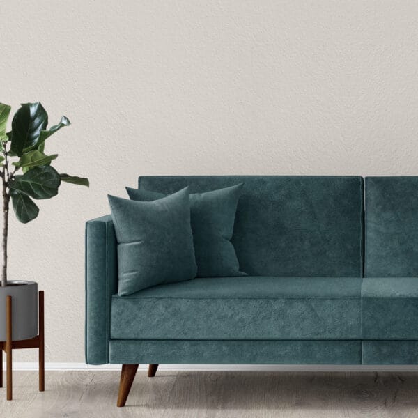 A modern living room with a teal velvet sofa adorned with matching cushions. A potted plant in a gray pot stands to the left. The room has a minimalist design with a light textured wall and wooden flooring.