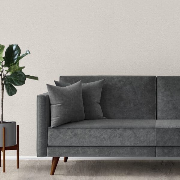 A modern living room with a gray velvet sofa featuring two matching pillows. A potted plant with lush green leaves is placed beside it on a wooden stand. The background shows a textured, light-colored wall.