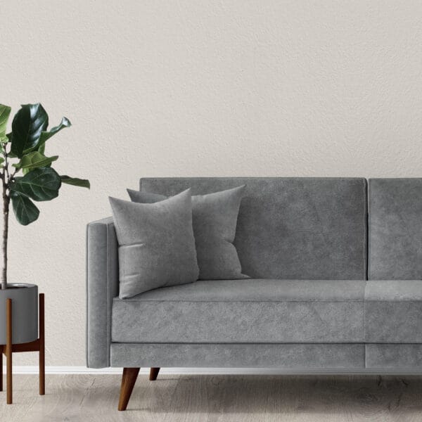 A modern living room with a gray sofa featuring two matching pillows, situated on a light wood floor. To the left of the sofa is a potted plant with green leaves in a sleek gray pot, in front of a neutral, textured wall.