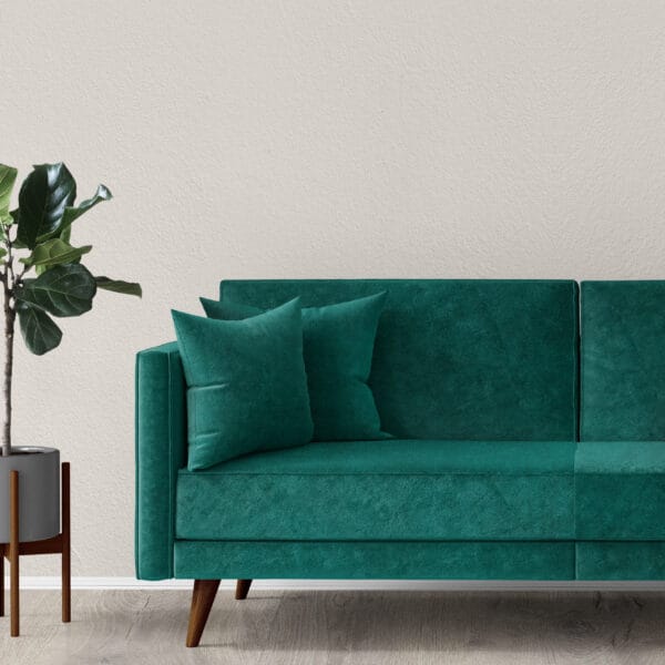 A modern living room featuring a green velvet sofa with two matching pillows, placed on a light wooden floor. A leafy plant in a gray pot stands beside the sofa against a textured beige wall. The scene is minimalistic and stylish.